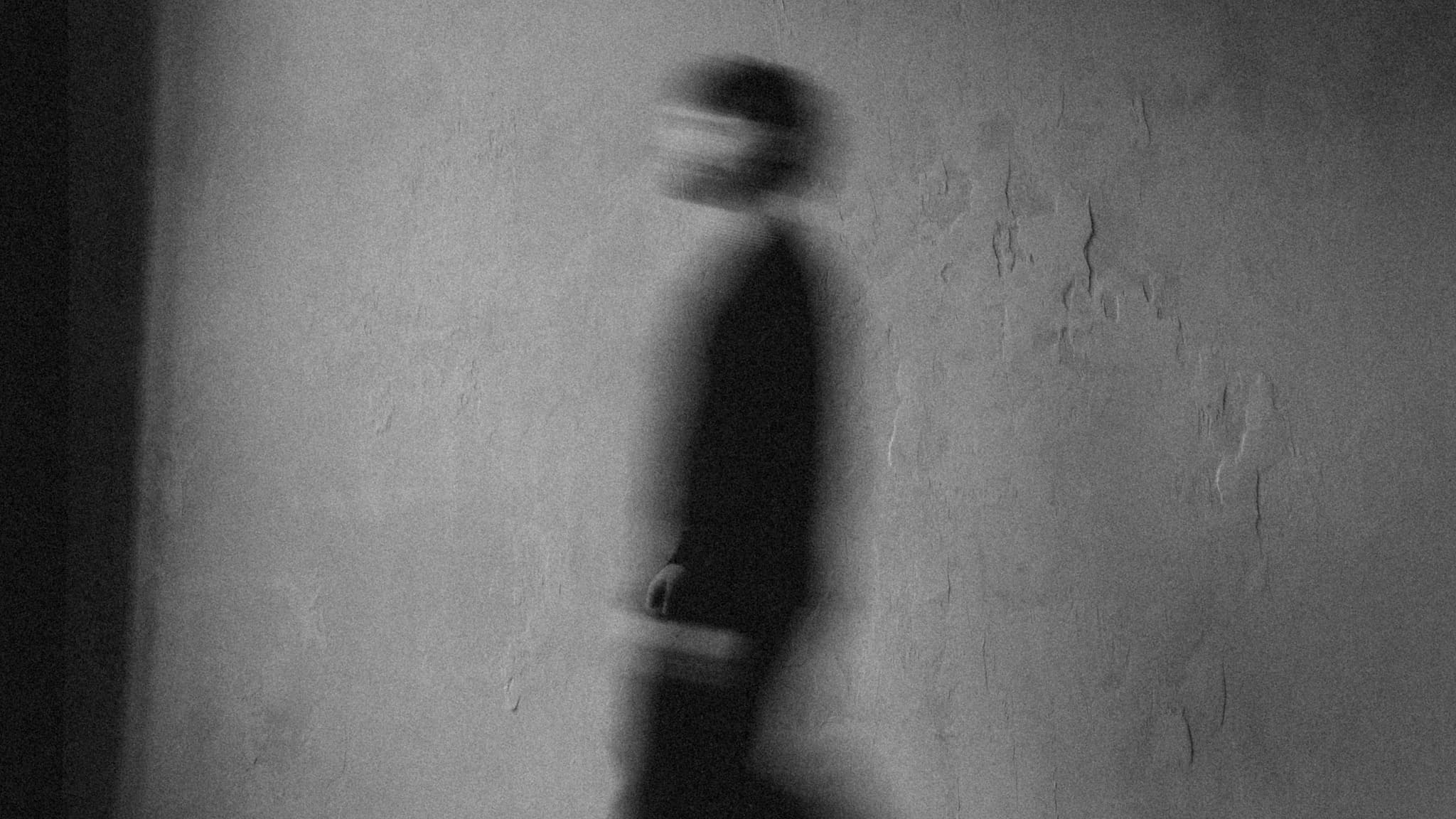 Long-exposure black and white photograph of a figure moving against a textured plaster wall