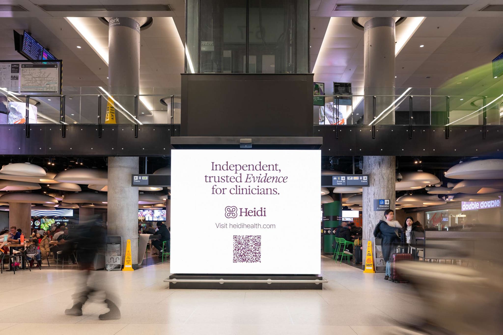 Freestanding Heidi screen reading “Independent, trusted Evidence for clinicians.” in a food court